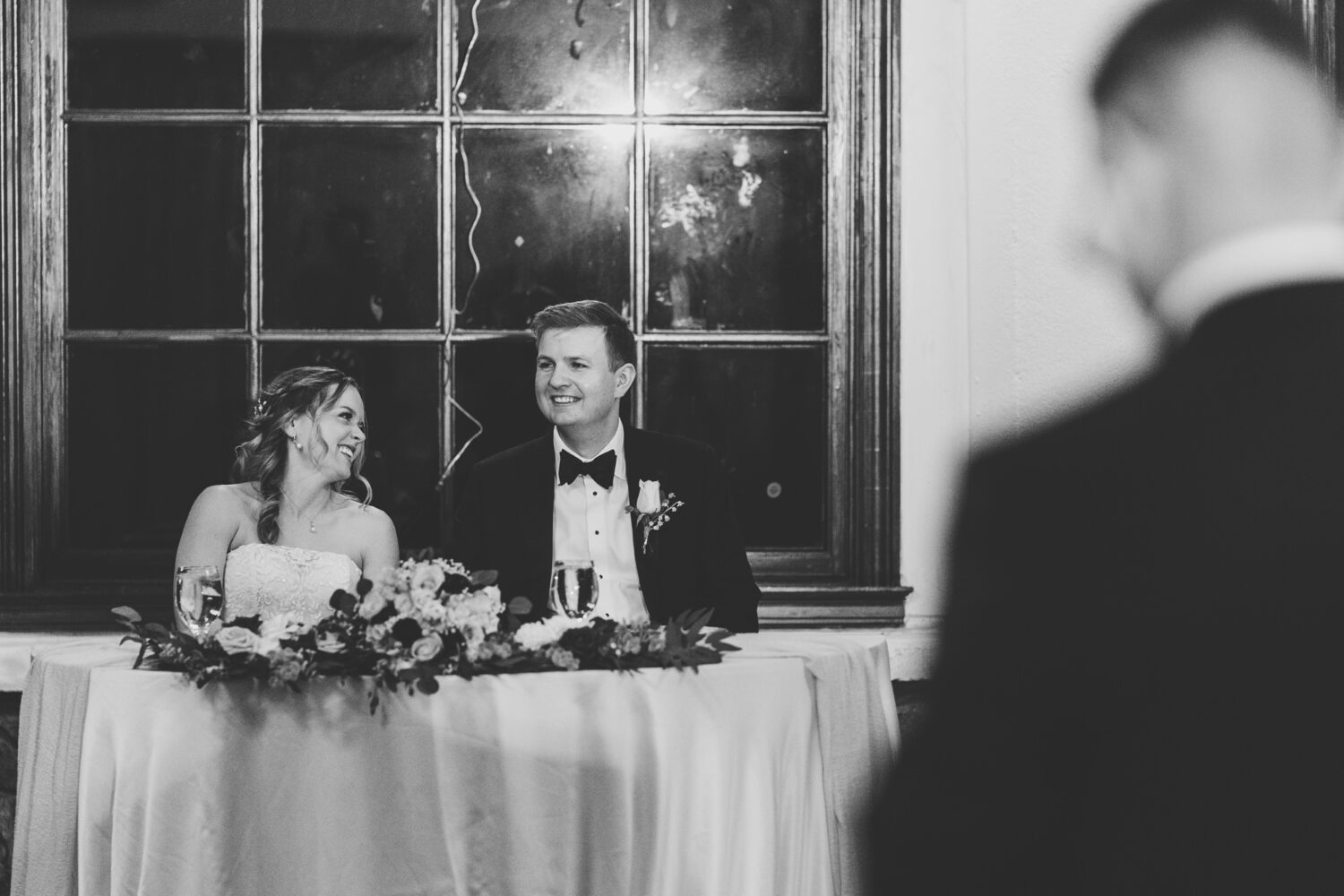 bride and groom laughing while listening to the bride's father giving a welcome speech