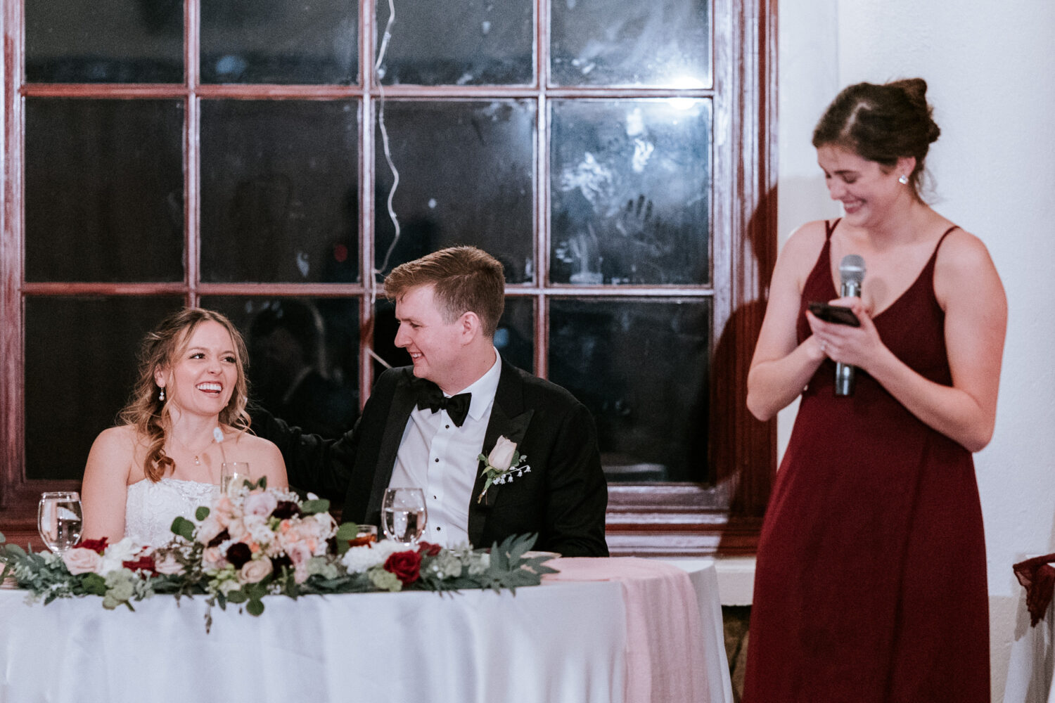 bride and groom laughing while listening to the matron of honor give her speech