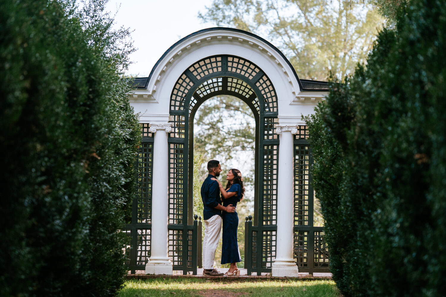 Couple standing under the white archway at Oatlands Historic House & Gardens in Leesburg, VA after their surprise proposal.